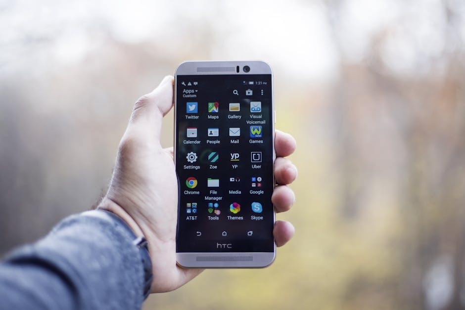 A close-up of a hand holding an HTC smartphone with visible apps on the screen, in an outdoor setting.