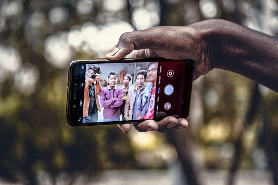 A group of friends captures a selfie outdoors using a smartphone in India. Close-up of hands holding the phone with faces in focus.