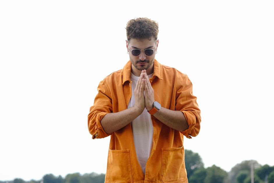 Stylish man in orange jacket posing outdoors with hands clasped.