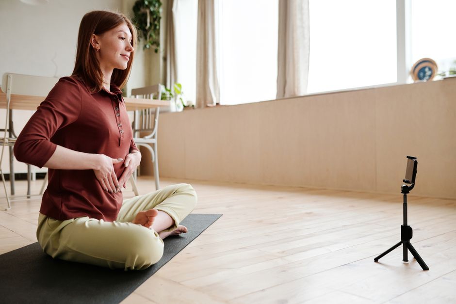 Young woman practicing yoga at home, recording a session on her smartphone with natural light streaming through windows.