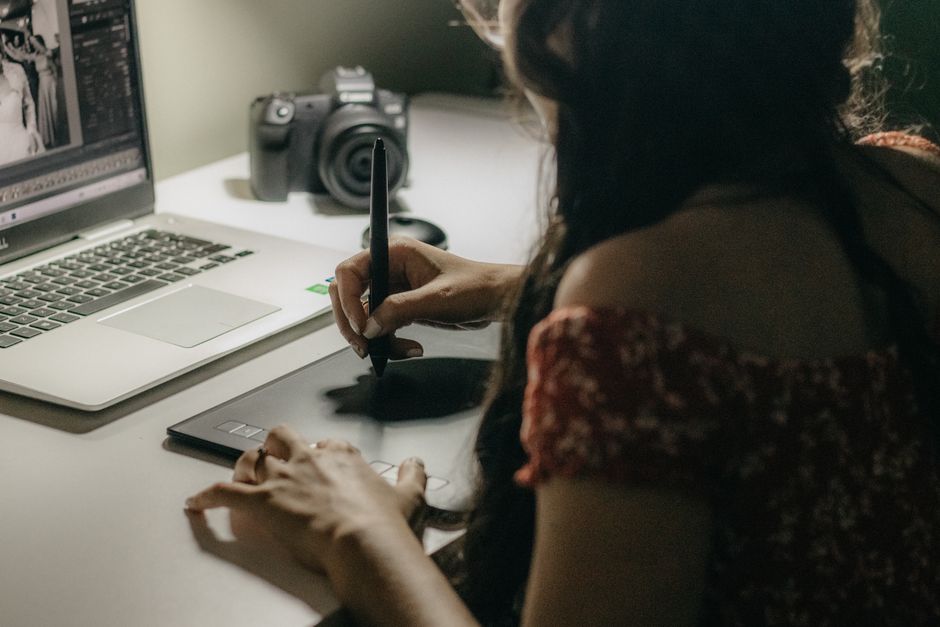 An adult woman using a drawing tablet while working on a laptop in a dimly lit room.